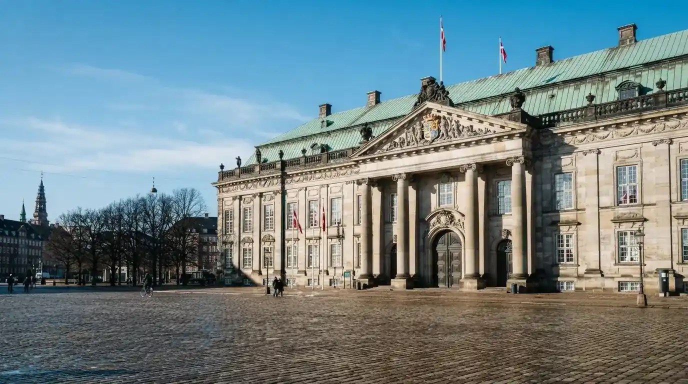 En historisk bygning i København med grøn kobbertag og kolonner i klart dagslys, set fra pladsen foran.